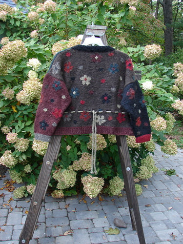 A Tara Handknit Sweater on a wooden stand, featuring colorful patterns and contrasting knit accents. Cozy ribbed neckline, cuffs, and hem. Oversized round metal cut buttons. Unique rear yarn gather tie. A luxurious and collectible piece. Bust 56, Waist 56, Hips 56, Length 21 inches.