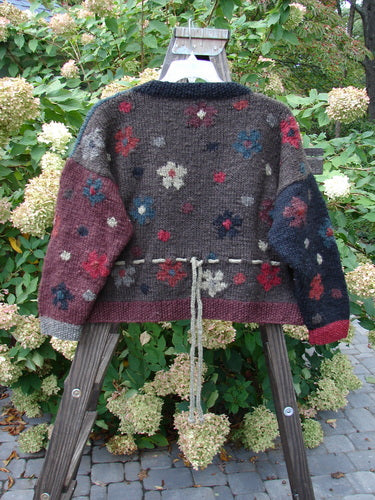 A Tara Handknit Sweater on a rack, featuring a cozy ribbed neckline, cuffs, and hem. Highly detailed in colorful patterns and contrasting knit accents. Oversized round metal cut buttons and a unique rear yarn gather tie. A luxurious, vintage beauty from the Hot Knots Sisters.