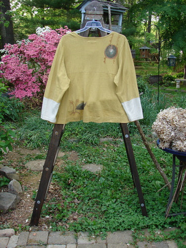 A close-up of a 2000 Cotton Hemp Philois Jacket in Dove and Gold, featuring a scooped varying hemline, contrasting sleeves, and taganut number buttons.