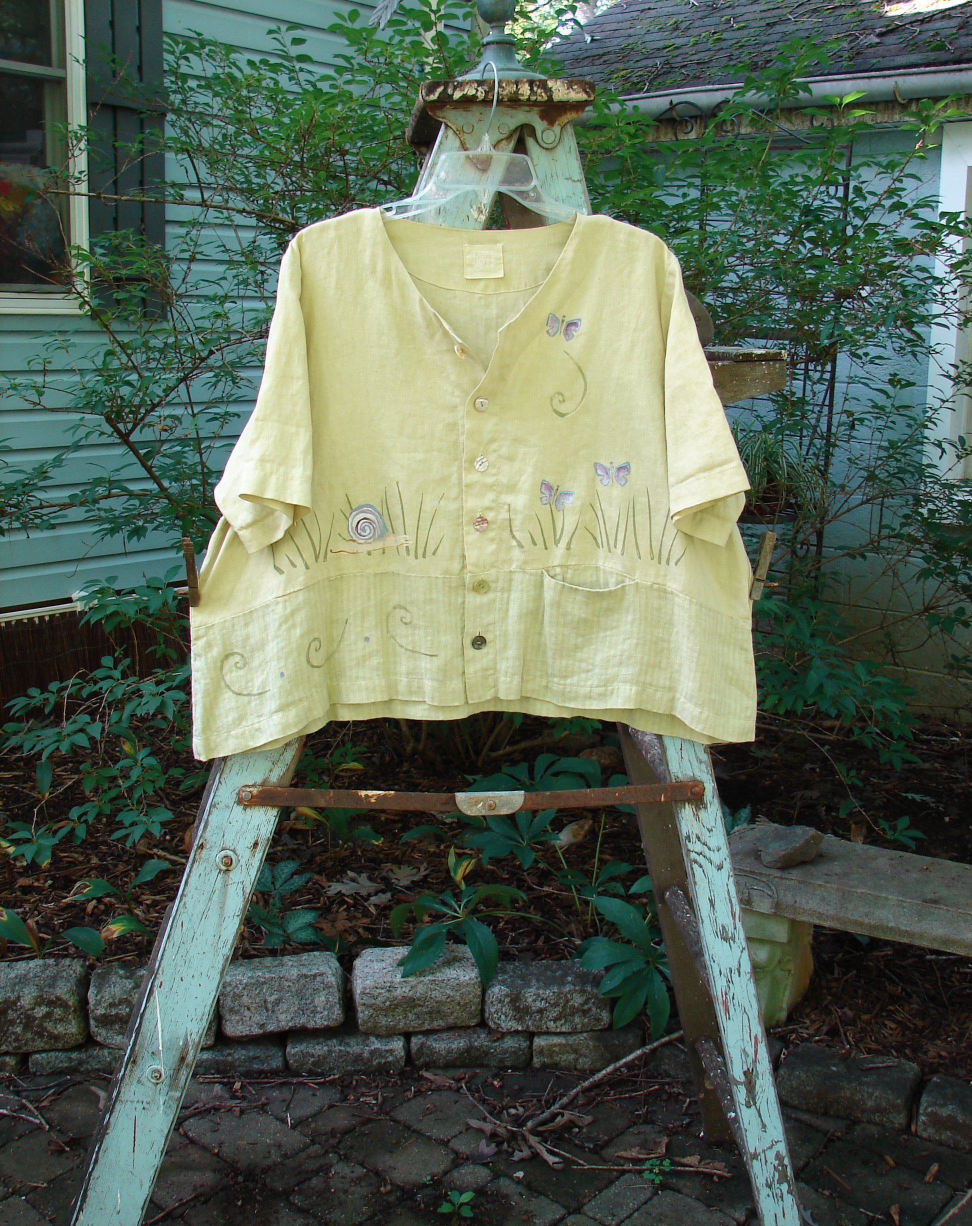1999 Cabana Top Butterfly Grass Citron Size 2: A yellow shirt with butterflies and snails on it, featuring a slight crop shape, shell buttons, and a varying hemline. Made from light textured linen, this top is simply flowing and light with a substantial weave. It also includes a double paneled contrasting fabric lower, a sweet drop pocket, and a signature Blue Fish patch and painted butterfly in the grass theme paint. Bust: 52, Waist: 54, Hips Hem Circumference: 60, Length: 23 inches.