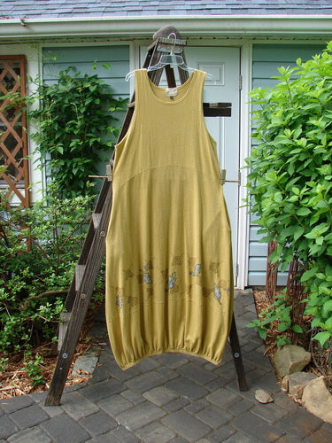 A dress on a ladder, featuring the 2000 Cotton Hemp Libby Jumper Fall Leaf Gold Size 1. The dress has a downward scooped waistline, 2 side pockets, and a bubbled shape. The jumper is made from a mid-weight hemp cotton knit fabric, providing a sophisticated drape and wondrous movement.