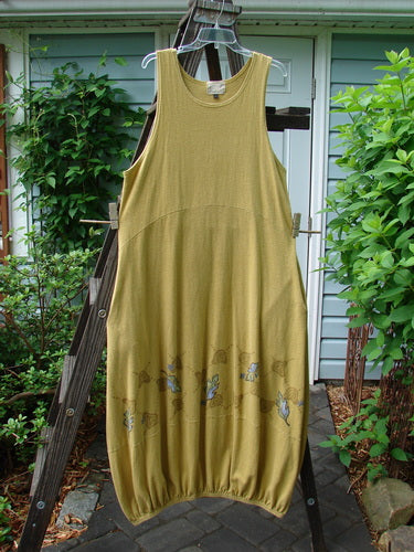 A yellow dress on a wooden rack, featuring a downward scooped waistline, 2 side pockets, and a bubbled shape. The Libby Jumper from the 2000 Cotton Hemp Resort Collection is made from a fabulous hemp cotton knit, offering a sophisticated drape and wondrous movement. The dress is adorned with a fall leaf theme paint, adding colorful and detailed accents. The slender size 1 measurements are as follows: Bust 42, Waist 44, Hips 48, and the length is 52 inches.