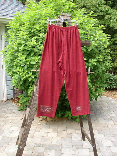 1997 Simple Pant Love Dove Regalia Size 1: A pair of red pants on a wooden rack, featuring a full elastic waistband, deep side pockets, and a slightly straighter fall. Made from organic cotton, these pants are part of the Spring Collection of 1997.