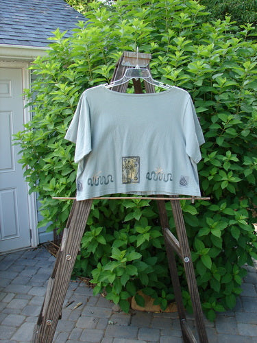 1993 Travel Top Many Moons Ocean Size 2: A t-shirt on a swinger with a wooden ladder under a bush.