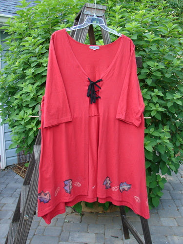 A red shirt with a black bow, featuring a Barclay Lace Up Panel Duster in Ruby. The shirt has a deep V-shaped neckline, flat tape lacings, a downward yoked rear drop panel, and widening three-quarter length sleeves. Bust: 50, Waist: 52, Hips: 54, Length: 43 inches. Made from organic cotton.