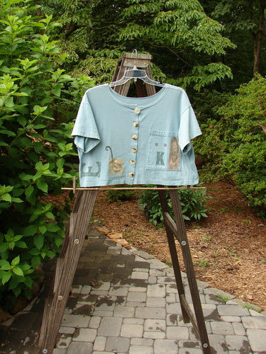 1994 Song Top Multi Letter "K" Ice Size 1: A shirt on a rack with a blue shirt on a wooden stand. Features a wide boxy shape, shallow neckline, vintage buttons, and a painted breast pocket.