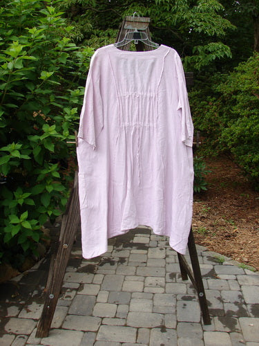 Image alt text: Barclay Linen Double Tie Back Jacket Sprig Pink Cloud Size 0 - A pink dress on a clothes rack with exterior vertical stitchery, front drop flop pockets, and a varying hemline.