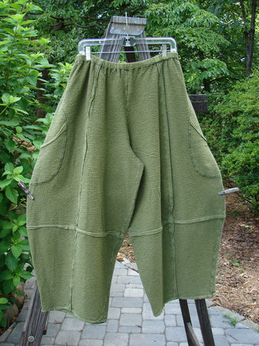A pair of Barclay Crepe Sectional Pocket 4 Square Pants in Olive, made from Heavy Weight Cotton Crepe. Features a unique bottom cut that drapes into a 3D diamond shape from the knee down. Complete with exterior wrap-around hip pockets, elastic waistband, and textured fabric. Size 1.