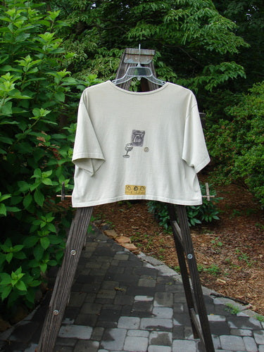 A white shirt with a vintage blue fish patch on a wooden rack. Oversized tee from the 1991 Summer Collection in wheat color.