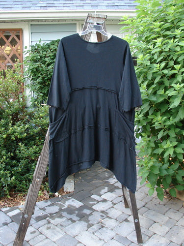 Barclay Reverse Stitch Double Pocket Tunic Dress: Black shirt on wooden rack. Mid-weight organic cotton. Lettuce-edged lower sleeves, banded and painted lower. Two drop front wrap-around exterior pockets. Three-quarter length sleeves. Continuous leaf garden theme paint. Size 2.