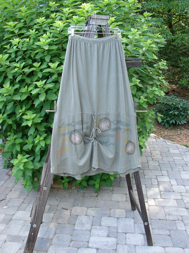 A 2000 Cotton Hemp Shade Skirt Biology Loden Size 2, hanging on a rack. The skirt features a full elastic waistband, an upward scoop, and varying hemline when tied with the extra long rippie. It has 3 vertical loops for manageable and creative adjustments, sectional horizontal panels, and the classic Resort Biology theme paint. Waist: 30-52, Hips: 52, Hem Circumference: 70, Length: 37 inches.