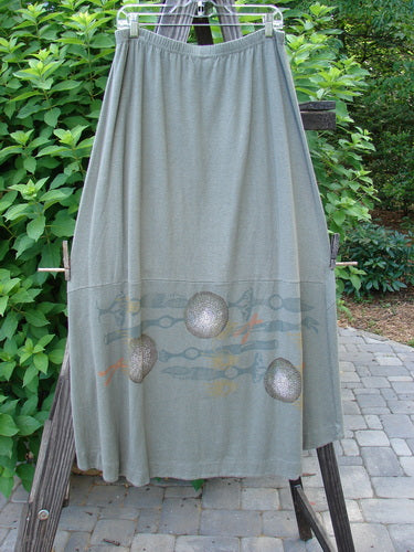 2000 Cotton Hemp Shade Skirt Biology Loden Size 2: A skirt on a rack, featuring a lovely hemp cotton blend fabric and a full elastic waistband. The skirt has an upward scoop and varying hemline when tied with the extra long rippie. It also has 3 vertical loops for adjustable styling and sectional horizontal panels. Perfect for the Resort Collection, this skirt showcases the classic Resort Biology theme paint.