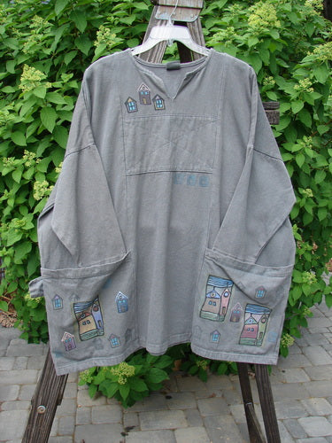 A grey shirt with a drawing on it, featuring a split front rolled neckline, oversized pockets, and village theme paint. Barclay Patched Canvas Urban Tunic Top Village Grey Stone Size 2.