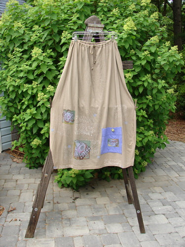 Barclay Big Pocket Skirt Shop Now Wheat Size 2: A skirt with a unique bell-shaped bottom, drawstring waistline, and giant exterior pocket with a vintage blue fish button closure. Two deep side pockets and a bottom swing add extra movement and flair.