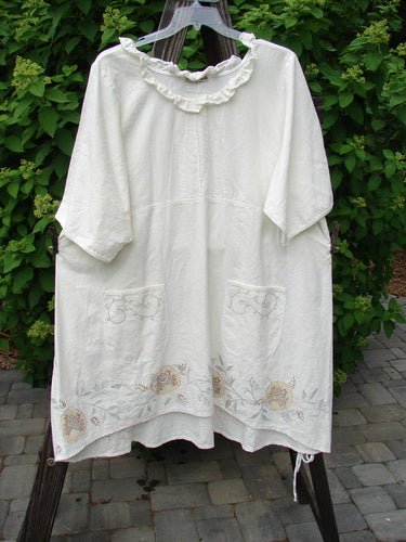 A white dress with a floral design on a clothes rack, featuring a sweet baby doll silhouette, thin pleats, varying hemline, and front painted pockets.