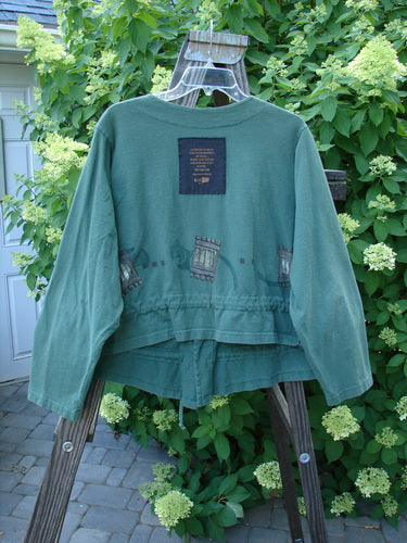 1997 Treehouse Jacket Many Windows Verdigris Size 2: A green jacket with a patch on it, made from mid-weight organic cotton. Features a double paneled V-neck, button line, and drawcord flounce.