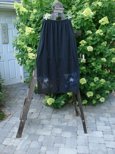 1997 Indra Skirt Florals Obsidian Size 2: A skirt on a wooden stand, featuring a full elastic waistline, double paneled lower, colorful chiseled buttons, deep side pockets, and whimsical floral theme paint.