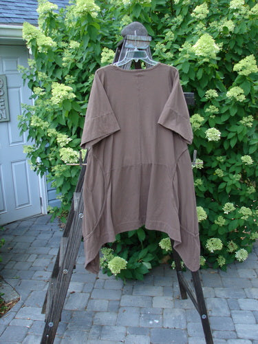 Image alt text: Barclay Double Pocket Bounce Tunic Dress, a brown shirt on a wooden rack, featuring wrap-around side curvy sectional seams, oversized front drop bushel pockets, and a dramatic upward scooped hemline.