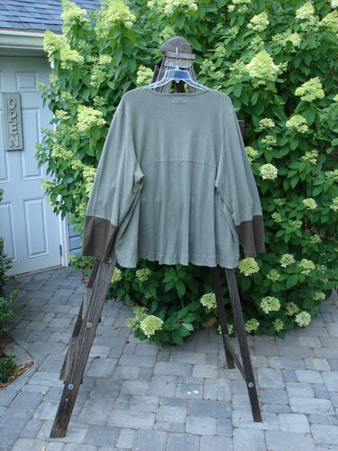 A long sleeved Philos Jacket in Loden on a wooden rack. Features include a scooped varying hemline, sectional horizontal panels, two lower pockets, taganut number buttons, and a rear flounce. Size 2, unpainted.