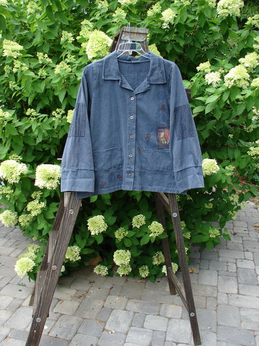 A blue jacket with patched stripe accents on a wooden ladder. The jacket features a full metal button front, double front drop rectangular thick patches, sectional panels, a stand-up collar, and an A-line shape. It has heavier double paneled cuffs and hemline, with super strip accents and a center rear patch. Size 1.
