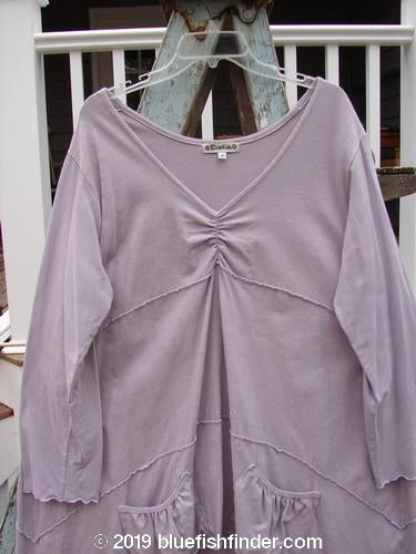 Barclay Cotton Lycra Vertical Gather Drop Pocket Dress Unpainted Lavender Size 2: A long-sleeved shirt on a swinger with a purple shirt on a white wooden rack.