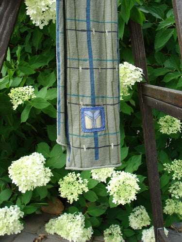 Image: A close-up of a towel on a wooden ladder with a close-up of a wooden chair and a close-up of a towel in the background. 

Alt text: Barclay Patched Scarf in Green Olive Plaid on a wooden ladder with a chair in the background.