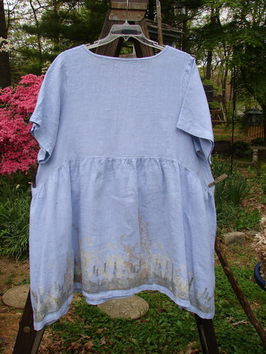 Barclay Linen Studio Pullover Dress Forest Border Sky Size 2: A blue shirt on a clothes rack with a close-up of the unique forest-themed border paint.
